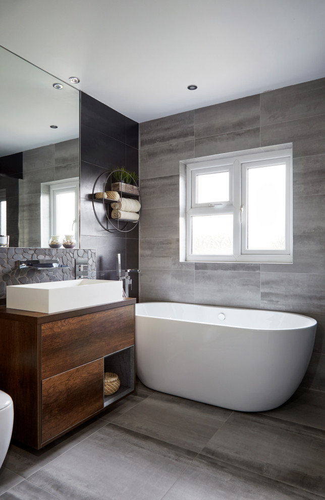 Modelo de cuarto de baño contemporáneo con armarios con paneles lisos, puertas de armario de madera en tonos medios, bañera exenta, paredes grises, lavabo sobreencimera y suelo gris