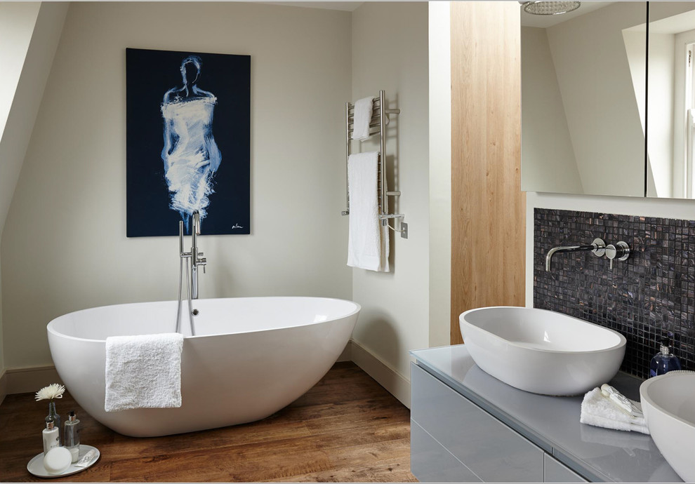 Idées déco pour une salle de bain contemporaine avec un placard à porte plane, des portes de placard grises, une baignoire indépendante, un carrelage noir, mosaïque, un mur gris, un sol en bois brun et une vasque.
