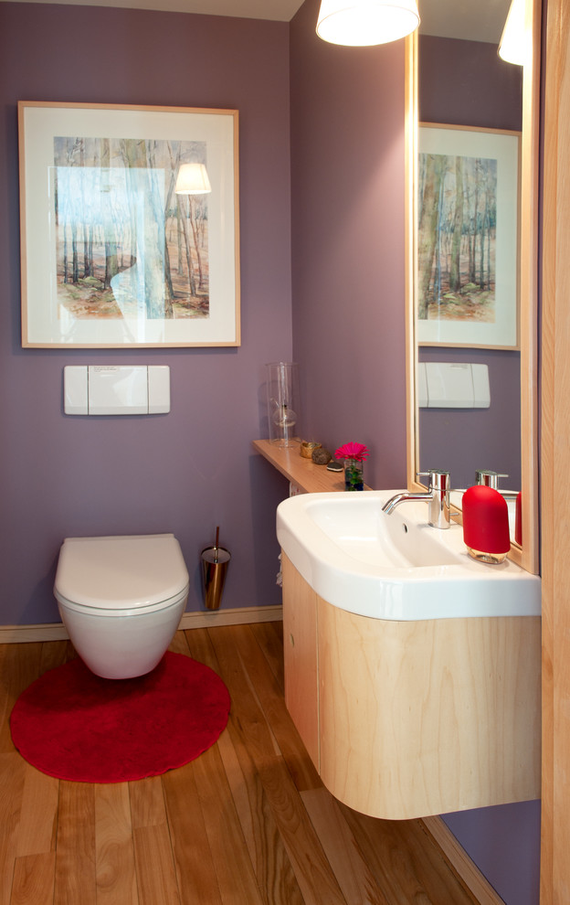 Imagen de cuarto de baño actual con puertas de armario de madera clara, sanitario de una pieza, paredes púrpuras, suelo de madera clara y encimera de acrílico
