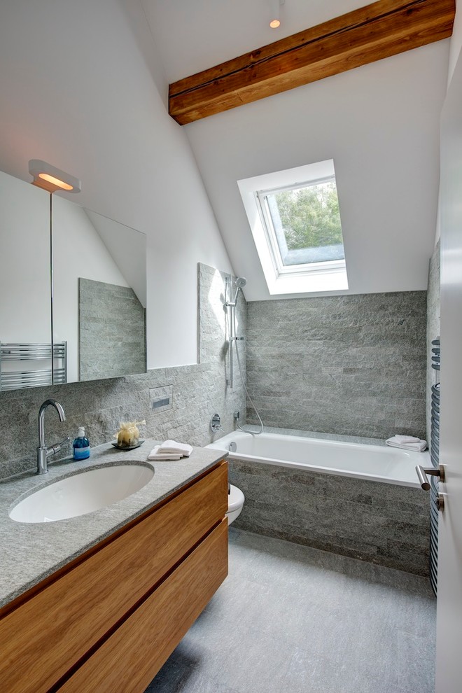Modern inredning av ett badrum, med ett undermonterad handfat, släta luckor, skåp i mellenmörkt trä, ett platsbyggt badkar, grå kakel och vita väggar