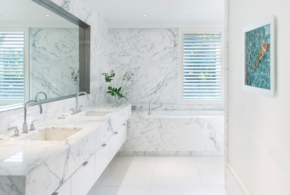 Inredning av ett modernt en-suite badrum, med ett integrerad handfat, släta luckor, vita skåp, ett undermonterat badkar, flerfärgad kakel och marmorkakel