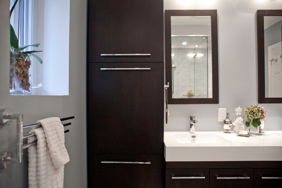 Modern inredning av ett litet badrum med dusch, med ett integrerad handfat, släta luckor, skåp i mörkt trä, bänkskiva i akrylsten, en hörndusch, en toalettstol med separat cisternkåpa, grå kakel, mosaik, grå väggar och klinkergolv i porslin