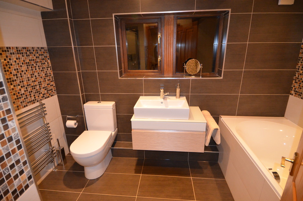 Photo of a medium sized contemporary ensuite bathroom in Gloucestershire with a wall-mounted sink, freestanding cabinets, light wood cabinets, glass worktops, a built-in bath, an alcove shower, a two-piece toilet, beige tiles, porcelain tiles, brown walls and porcelain flooring.