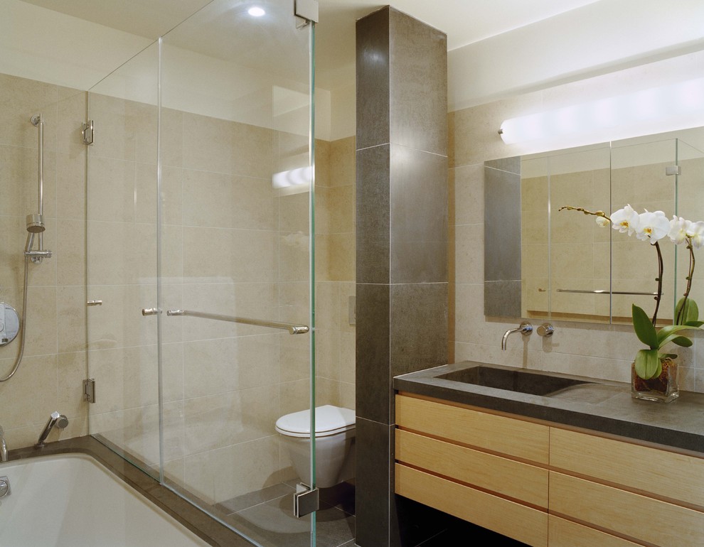 This is an example of a large contemporary ensuite bathroom in New York with flat-panel cabinets, light wood cabinets, a built-in bath, a shower/bath combination, a wall mounted toilet, beige tiles, ceramic tiles, beige walls, porcelain flooring, an integrated sink, limestone worktops, grey floors, a sliding door and grey worktops.