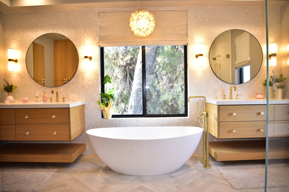 Cette image montre une salle de bain principale et beige et blanche design en bois brun avec une baignoire indépendante, un lavabo intégré, un plan de toilette blanc, un placard à porte plane, un carrelage beige, un mur beige, parquet clair et un sol beige.
