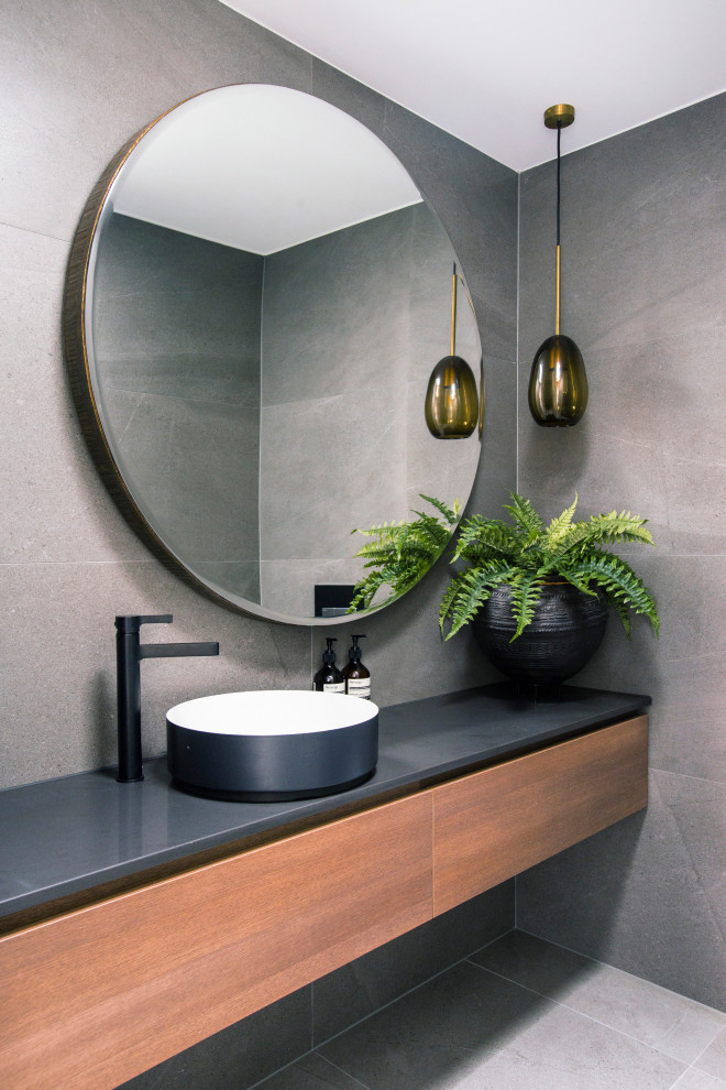 Foto på ett funkis grå badrum, med släta luckor, skåp i mellenmörkt trä, grå kakel, ett fristående handfat och grått golv