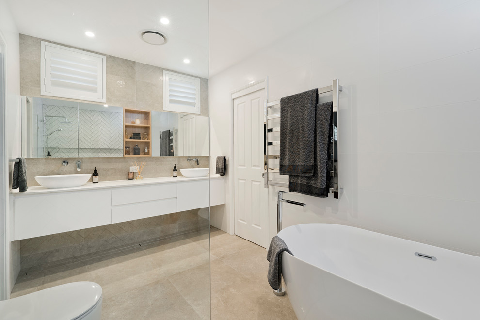 Inspiration for a medium sized contemporary ensuite bathroom in Brisbane with flat-panel cabinets, white cabinets, a freestanding bath, a walk-in shower, a wall mounted toilet, porcelain tiles, white walls, porcelain flooring, a vessel sink, engineered stone worktops, beige floors, an open shower, white worktops and grey tiles.