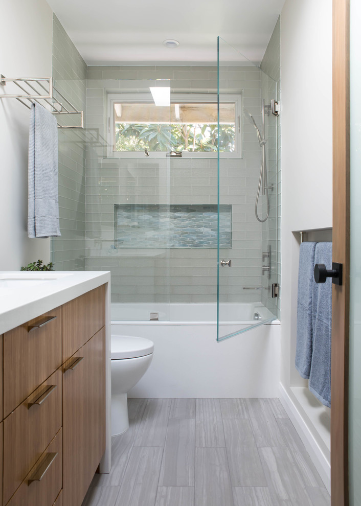 This is an example of a contemporary shower room bathroom in Los Angeles with flat-panel cabinets, medium wood cabinets, an alcove bath, a shower/bath combination, a one-piece toilet, glass tiles, porcelain flooring, a submerged sink, engineered stone worktops, grey floors, a hinged door and white worktops.