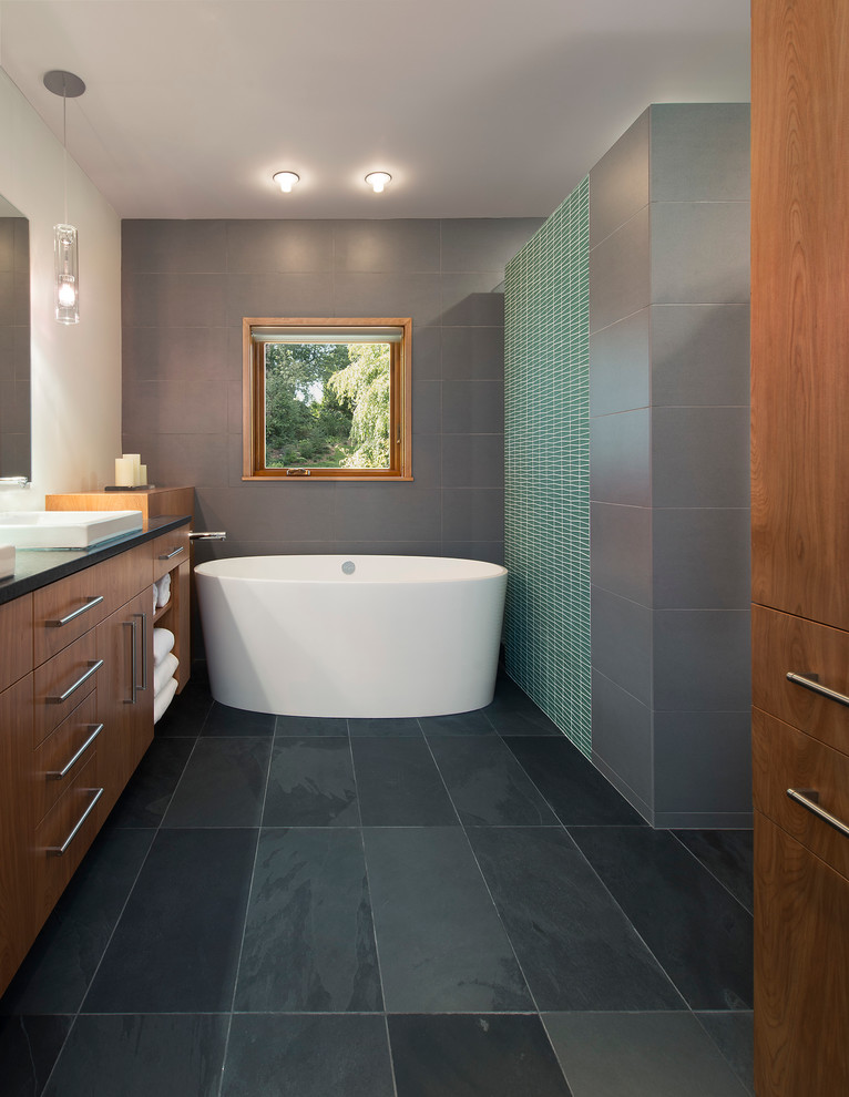 Example of a large trendy master gray tile and porcelain tile slate floor freestanding bathtub design in DC Metro with a drop-in sink, flat-panel cabinets, medium tone wood cabinets, granite countertops and gray walls