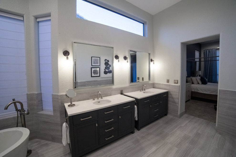 Cette photo montre une salle de bain principale chic avec un placard à porte shaker, des portes de placard noires, une baignoire indépendante, un carrelage gris, des carreaux de céramique, un mur blanc, un sol en vinyl, un lavabo encastré et un plan de toilette en surface solide.