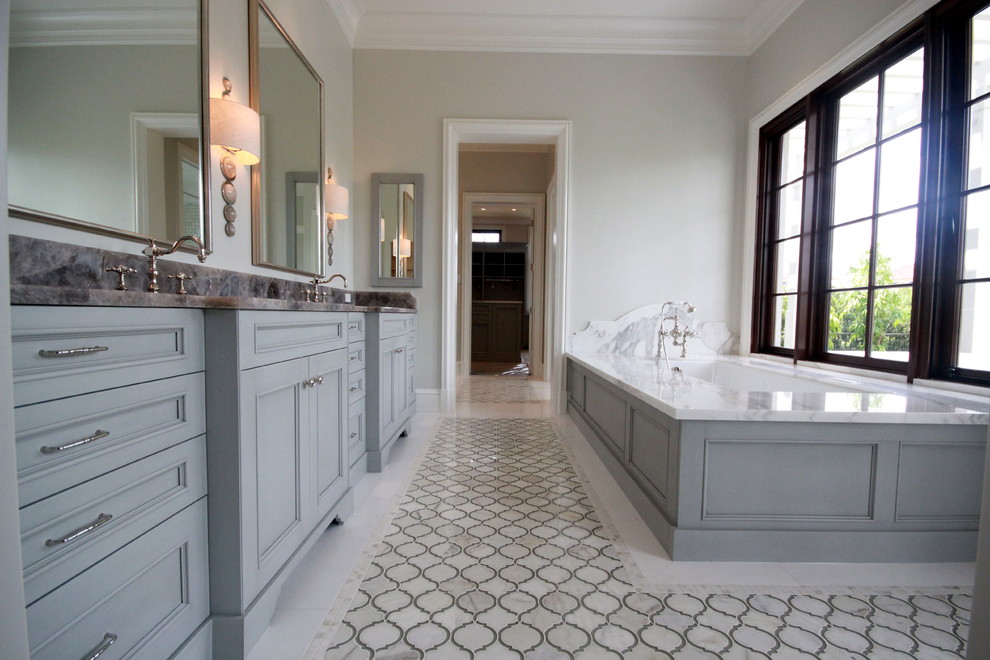 Cette image montre une salle de bain principale et grise et blanche traditionnelle de taille moyenne avec un placard avec porte à panneau encastré, des portes de placard grises, une baignoire encastrée, un mur gris, un lavabo encastré, un carrelage blanc, du carrelage en marbre, un sol en carrelage de céramique, un plan de toilette en marbre et un sol beige.