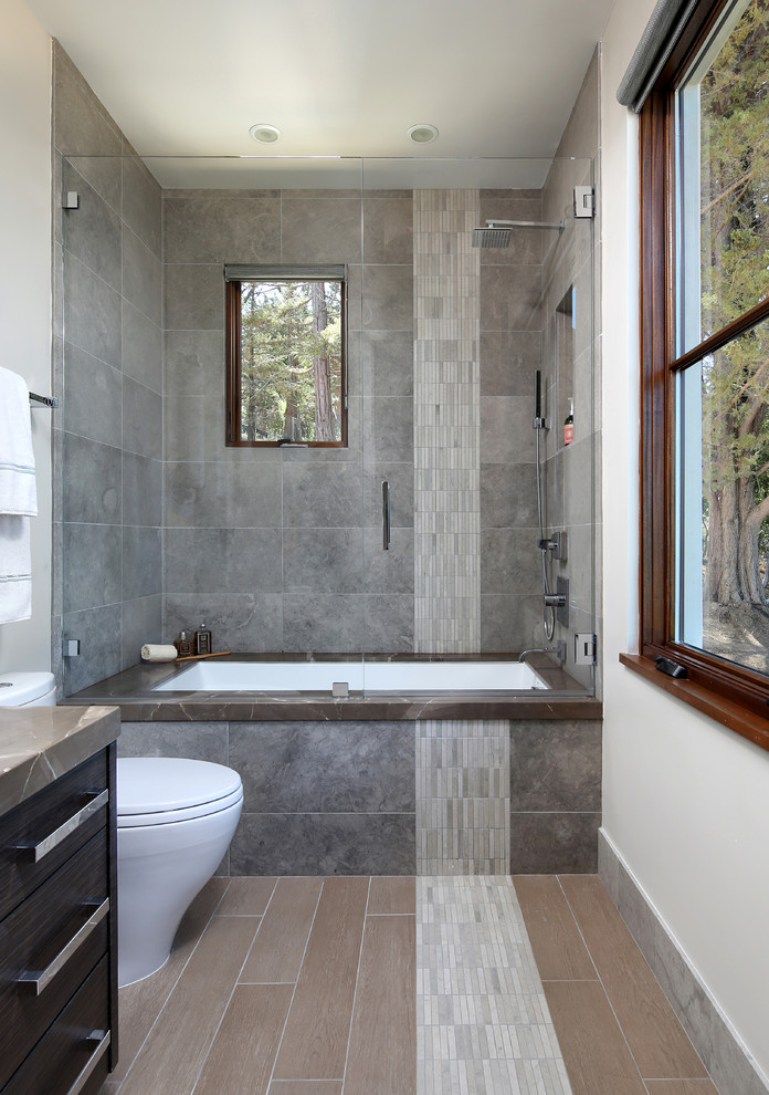 Inspiration pour une salle de bain design en bois foncé avec un placard à porte plane, une baignoire encastrée, un combiné douche/baignoire, un carrelage gris, un mur blanc, WC à poser et une cabine de douche à porte battante.