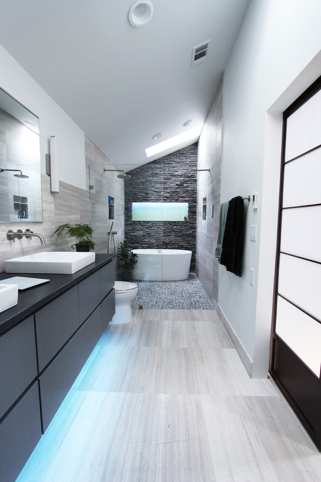 Example of a mid-sized trendy master gray tile and stone tile limestone floor, gray floor, double-sink and vaulted ceiling bathroom design in Atlanta with flat-panel cabinets, gray cabinets, white walls, a two-piece toilet, a vessel sink, granite countertops, black countertops, a niche and a floating vanity