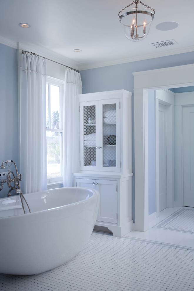 Modelo de cuarto de baño principal costero con armarios estilo shaker, puertas de armario blancas, bañera exenta, paredes azules, suelo con mosaicos de baldosas, lavabo bajoencimera y encimera de mármol