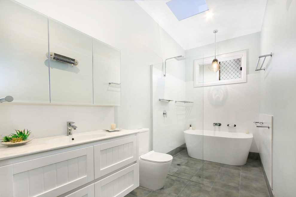 Пример оригинального дизайна: большая главная ванная комната в стиле неоклассика (современная классика) с белыми фасадами, отдельно стоящей ванной, душем без бортиков, белой плиткой, белыми стенами, монолитной раковиной, серым полом, открытым душем, белой столешницей, унитазом-моноблоком, полом из керамической плитки и фасадами с утопленной филенкой
