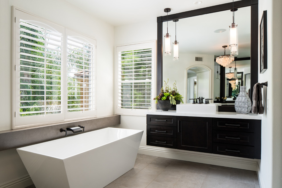 На фото: главная ванная комната в стиле неоклассика (современная классика) с фасадами в стиле шейкер, темными деревянными фасадами, отдельно стоящей ванной, белыми стенами, настольной раковиной, серым полом и зеркалом с подсветкой с