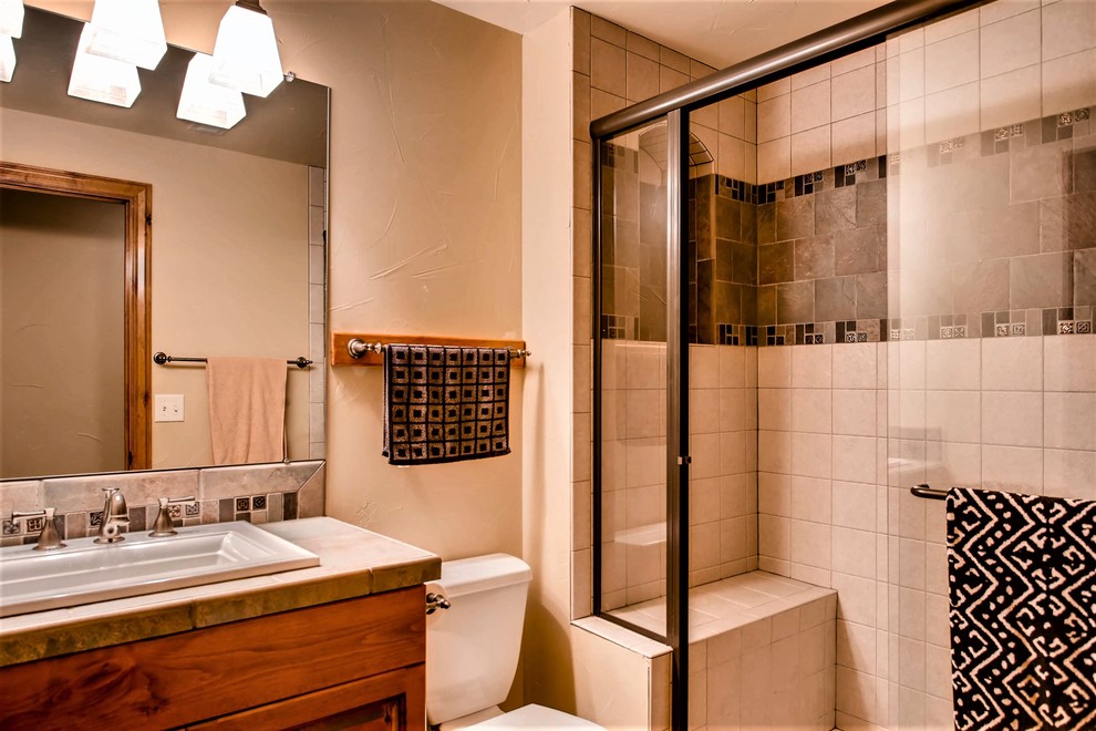 Exempel på ett mellanstort amerikanskt beige beige badrum med dusch, med luckor med upphöjd panel, skåp i mellenmörkt trä, en dusch i en alkov, en toalettstol med separat cisternkåpa, beige kakel, beige väggar, ett nedsänkt handfat, kaklad bänkskiva, dusch med skjutdörr och keramikplattor