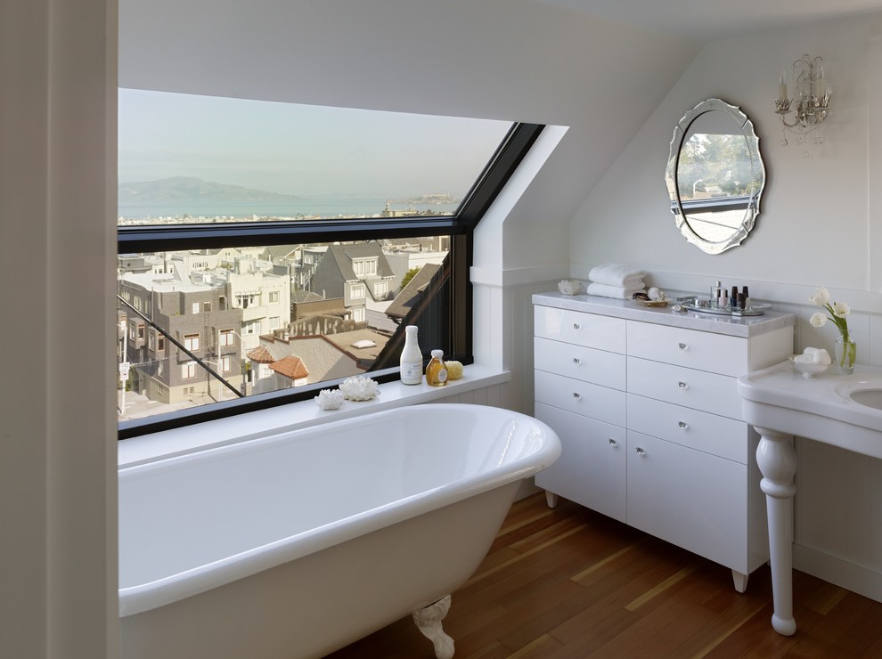 Inredning av ett klassiskt badrum, med ett konsol handfat, släta luckor, vita skåp, ett badkar med tassar, vita väggar och mellanmörkt trägolv