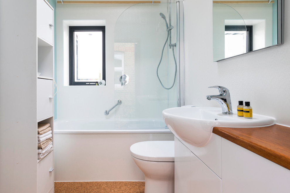 Exempel på ett litet modernt brun brunt badrum för barn, med släta luckor, vita skåp, ett badkar i en alkov, en dusch i en alkov, en toalettstol med hel cisternkåpa, glasskiva, vita väggar, korkgolv, ett nedsänkt handfat, träbänkskiva, brunt golv och dusch med gångjärnsdörr