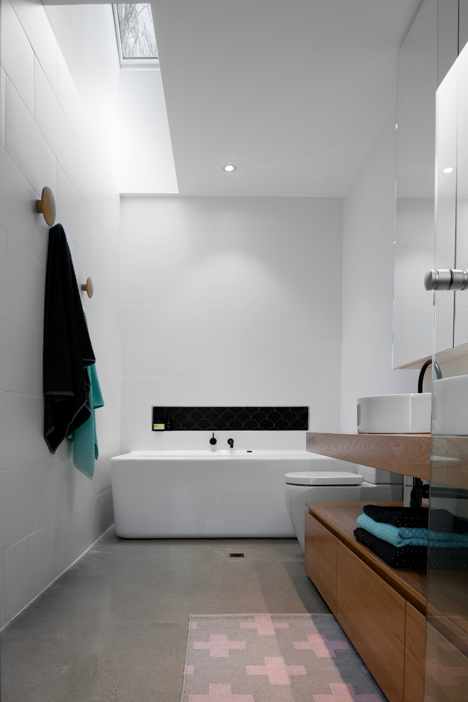 Nordisches Badezimmer mit verzierten Schränken, freistehender Badewanne, Toilette mit Aufsatzspülkasten, Porzellanfliesen, weißer Wandfarbe, Betonboden, Waschtischkonsole und Waschtisch aus Holz in Adelaide