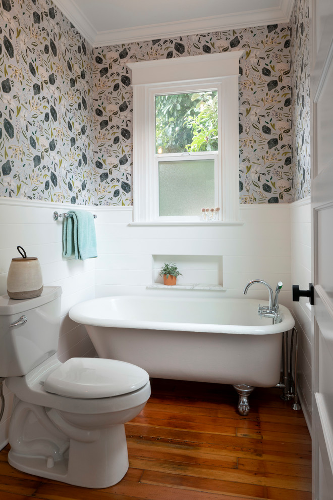Bild på ett litet amerikanskt badrum, med ett badkar med tassar, en dusch/badkar-kombination, en toalettstol med separat cisternkåpa, vit kakel, keramikplattor, vita väggar, ljust trägolv, ett väggmonterat handfat och dusch med duschdraperi