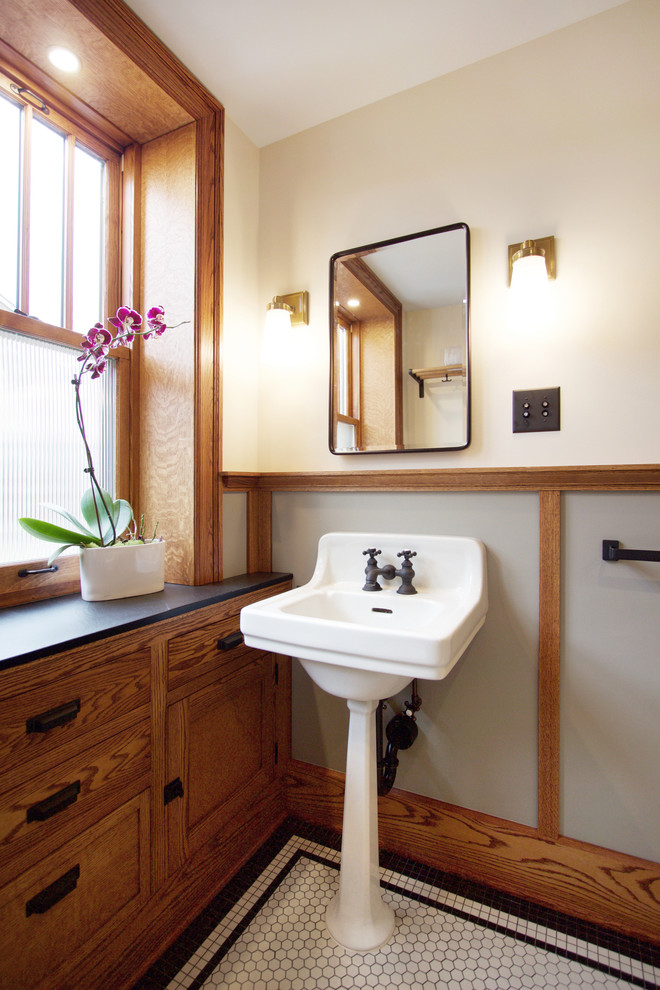 Modelo de cuarto de baño de estilo americano con armarios estilo shaker, puertas de armario de madera oscura, suelo con mosaicos de baldosas, lavabo con pedestal, suelo blanco y encimeras negras