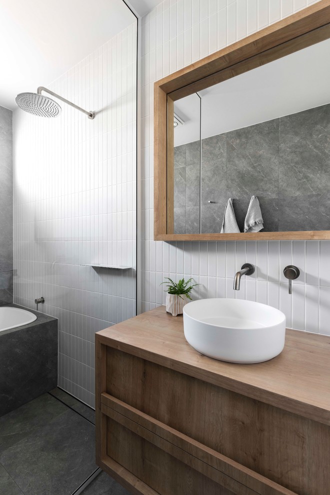 Bild på ett funkis brun brunt badrum, med ett platsbyggt badkar, ett fristående handfat, träbänkskiva, med dusch som är öppen, luckor med infälld panel, skåp i mellenmörkt trä, våtrum, vit kakel och grått golv