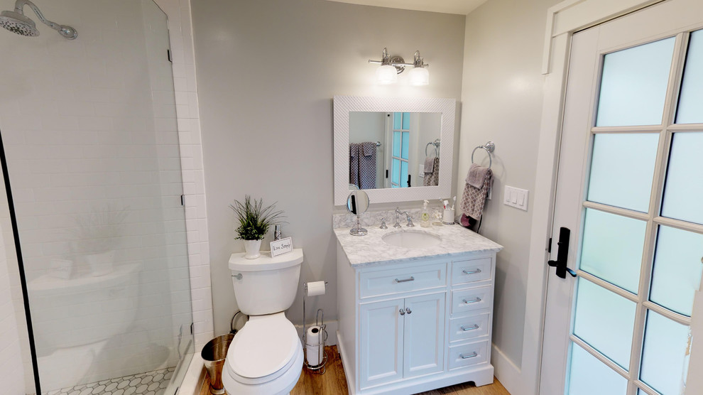 Corner shower - small traditional 3/4 white tile and subway tile medium tone wood floor corner shower idea in Los Angeles with recessed-panel cabinets, white cabinets, a two-piece toilet, white walls, an undermount sink, quartz countertops, a hinged shower door and multicolored countertops