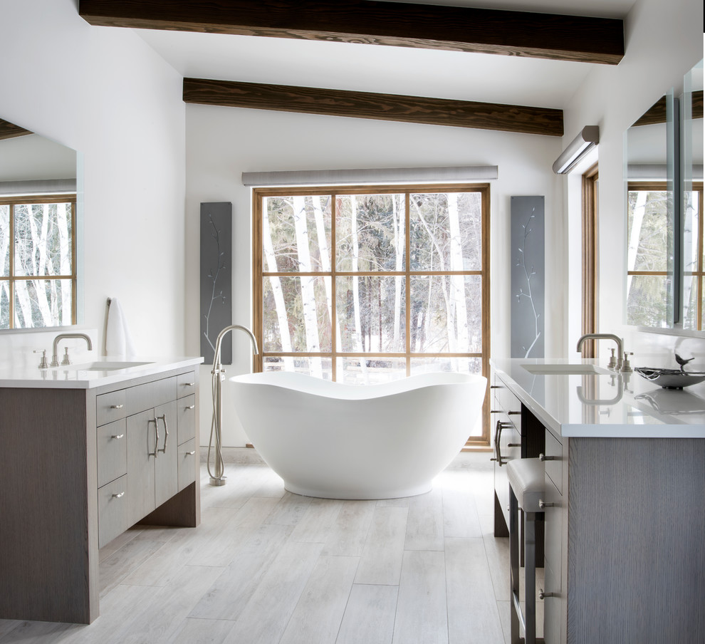 Cette image montre une salle de bain principale chalet avec un placard à porte plane, des portes de placard grises, une baignoire indépendante, un mur blanc, un lavabo encastré, un sol beige et un plan de toilette blanc.