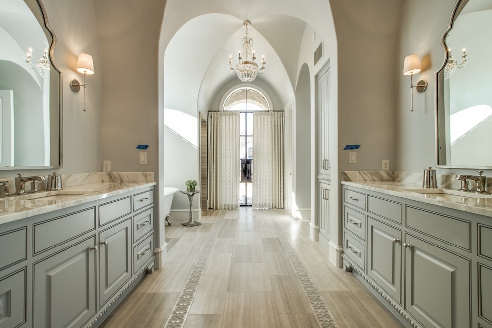 Réalisation d'une très grande salle de bain principale tradition avec des portes de placard grises, une baignoire sur pieds, un mur beige, un lavabo encastré, un sol beige, un placard à porte affleurante, un sol en carrelage de porcelaine et un plan de toilette en marbre.