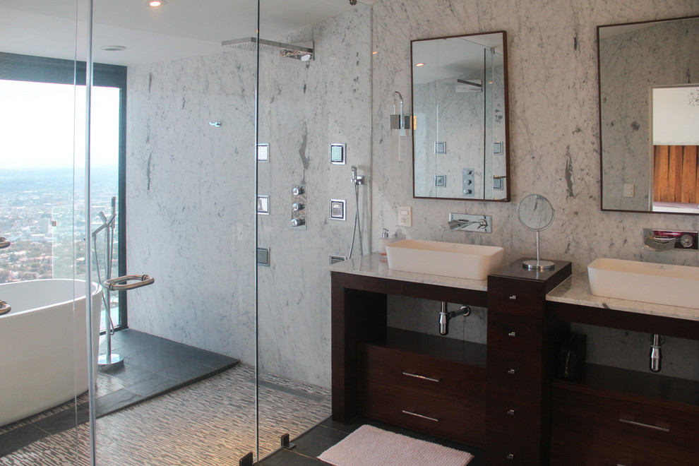 На фото: огромная главная ванная комната в современном стиле с врезной раковиной, фасадами островного типа, темными деревянными фасадами, мраморной столешницей, двойным душем, инсталляцией, черной плиткой, каменной плиткой, белыми стенами, полом из сланца и отдельно стоящей ванной с