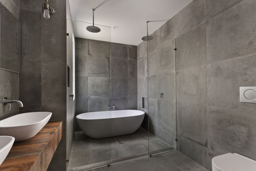 Industriell inredning av ett stort brun brunt en-suite badrum, med ett fristående badkar, en dubbeldusch, grå kakel, grå väggar, betonggolv, ett fristående handfat, träbänkskiva, öppna hyllor, skåp i mellenmörkt trä och en vägghängd toalettstol