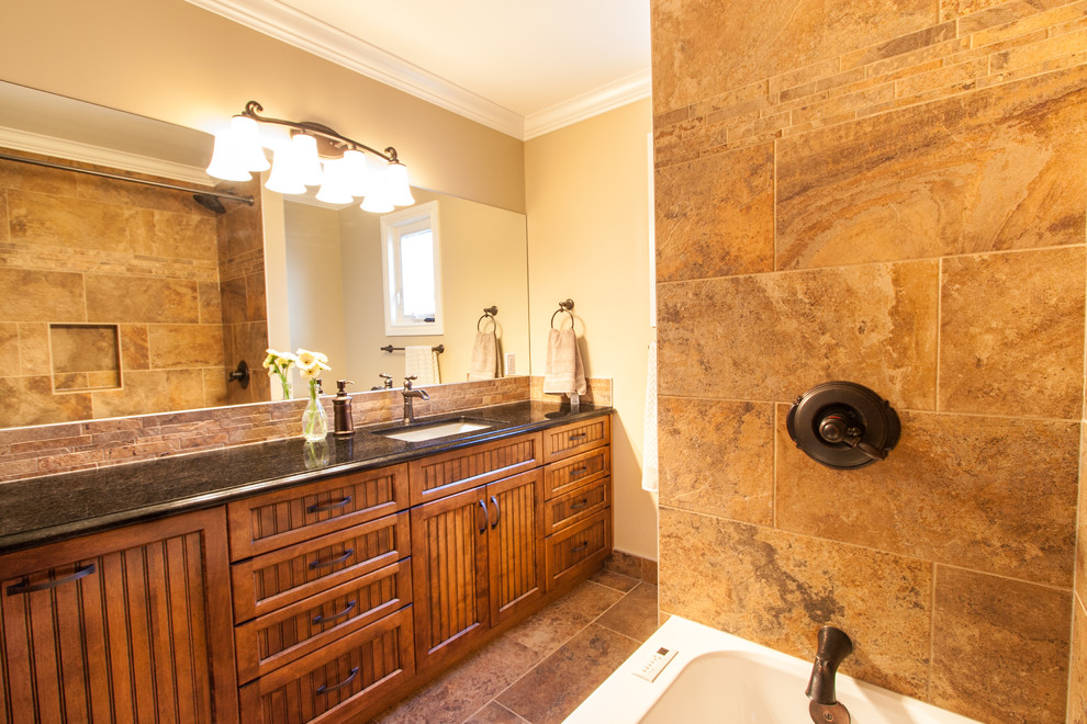 Klassisk inredning av ett litet badrum, med möbel-liknande, bruna skåp, granitbänkskiva, ett badkar i en alkov, en dusch/badkar-kombination, brun kakel, stenkakel, bruna väggar, klinkergolv i keramik och ett undermonterad handfat