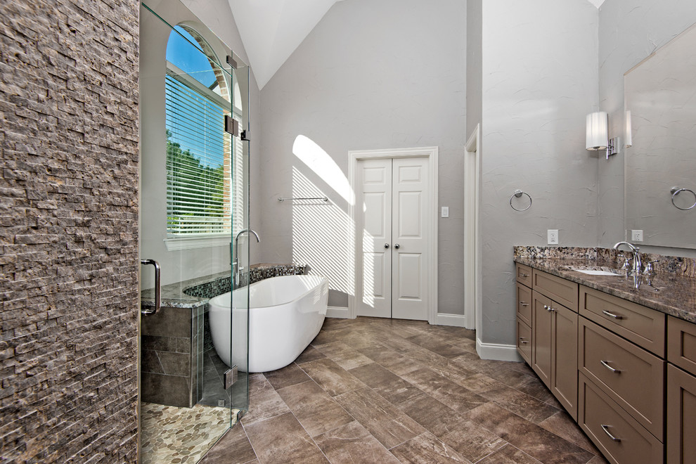 Klassisk inredning av ett en-suite badrum, med luckor med infälld panel, bruna skåp, ett fristående badkar, en kantlös dusch, brun kakel, porslinskakel, grå väggar, klinkergolv i porslin, granitbänkskiva och ett undermonterad handfat