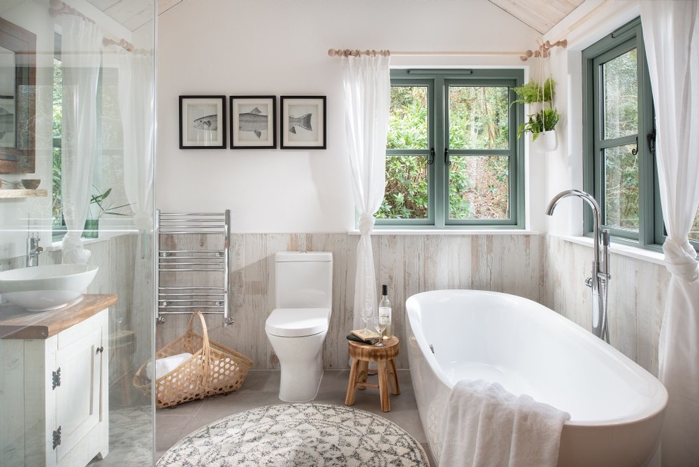 This is an example of a country bathroom in Cornwall with shaker cabinets, white cabinets, a freestanding bath, a one-piece toilet, white walls, a vessel sink, wooden worktops, grey floors and brown worktops.