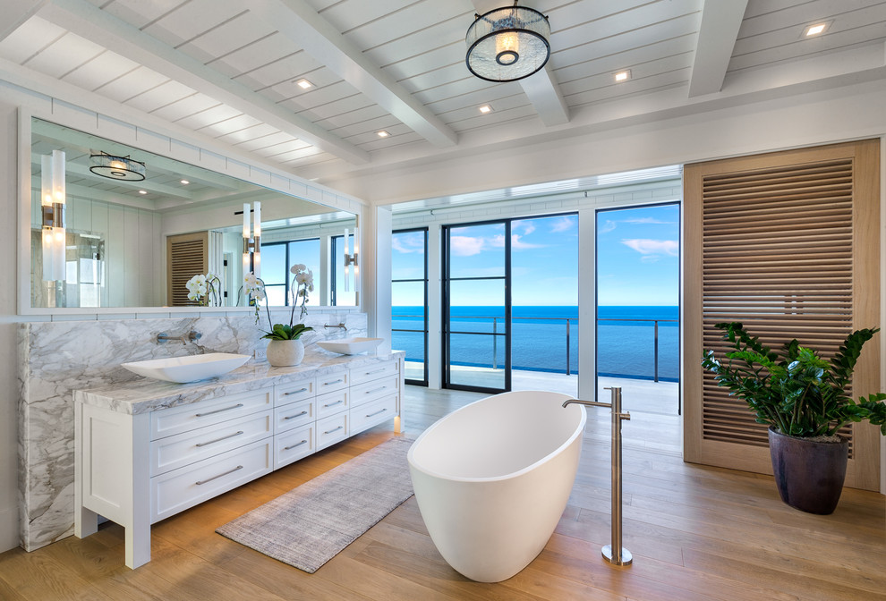 Example of a beach style medium tone wood floor and brown floor freestanding bathtub design in Orange County with shaker cabinets, white cabinets, white walls, a vessel sink and gray countertops