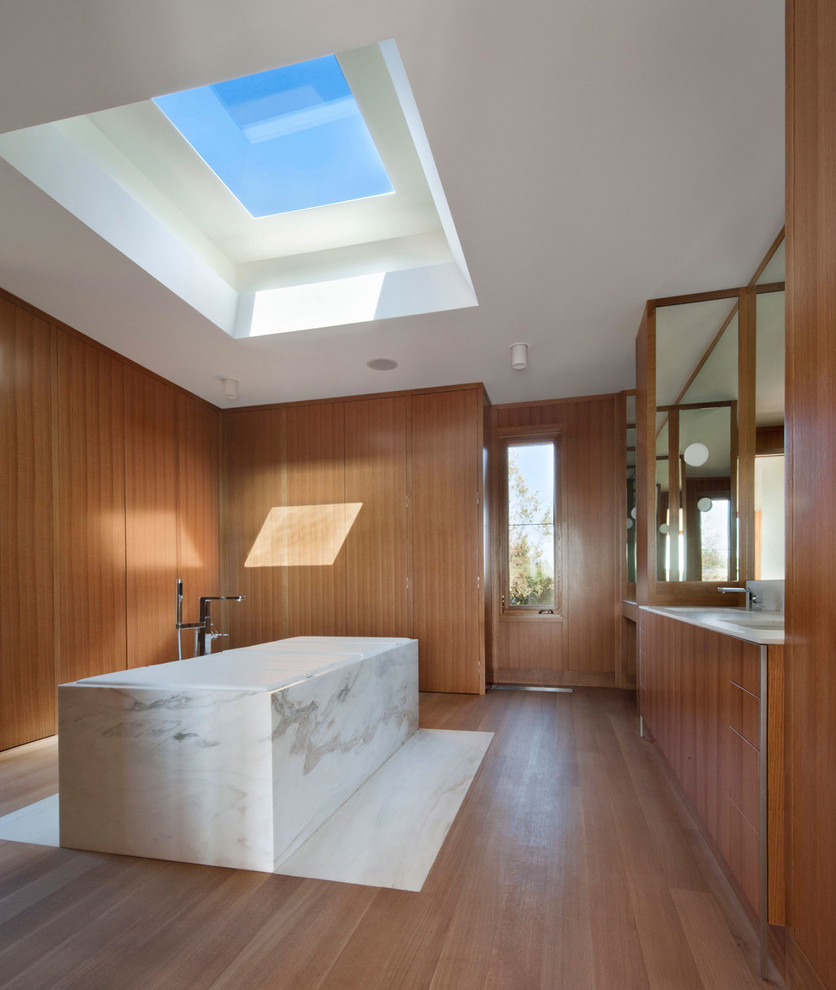 Cette photo montre une grande salle de bain principale moderne en bois brun avec un placard à porte plane, une baignoire indépendante, un sol en bois brun, un plan de toilette en marbre, un carrelage blanc, un mur marron, un lavabo encastré, un sol marron et un plan de toilette blanc.
