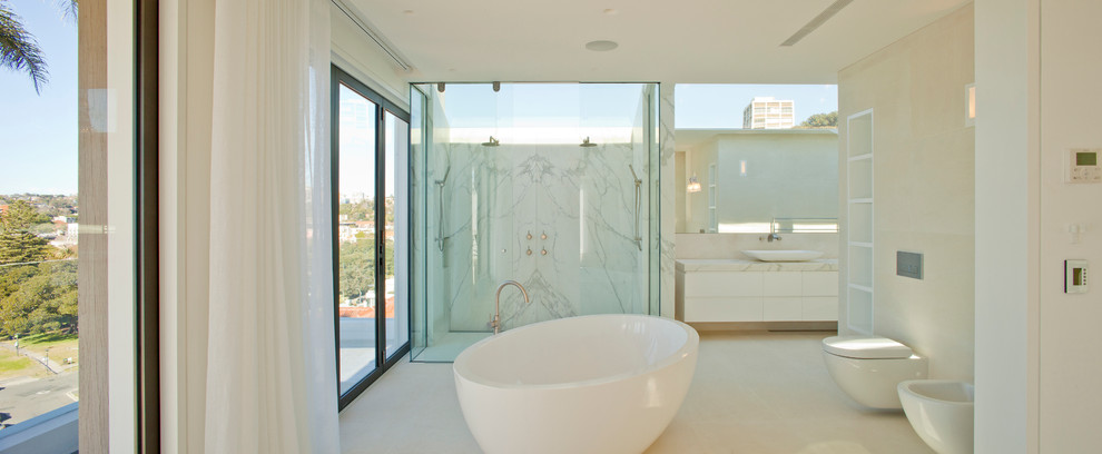 This is an example of a large mediterranean ensuite bathroom in Sydney with a vessel sink, white cabinets, limestone worktops, a freestanding bath, a double shower, a wall mounted toilet, white tiles, stone tiles, white walls and limestone flooring.