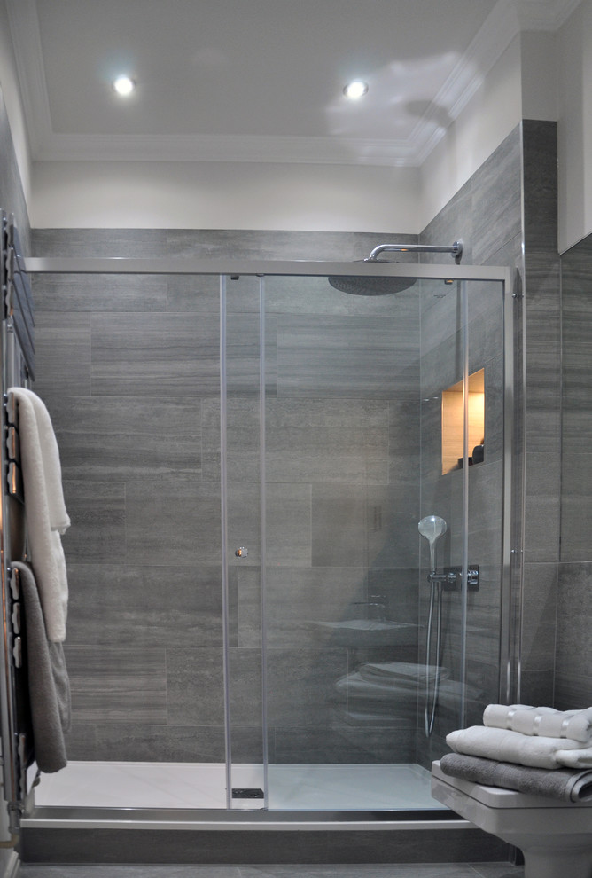 This is an example of a medium sized contemporary ensuite bathroom in London with a built-in sink, flat-panel cabinets, grey cabinets, solid surface worktops, a built-in bath, a shower/bath combination, a wall mounted toilet, beige tiles, ceramic tiles, white walls and ceramic flooring.