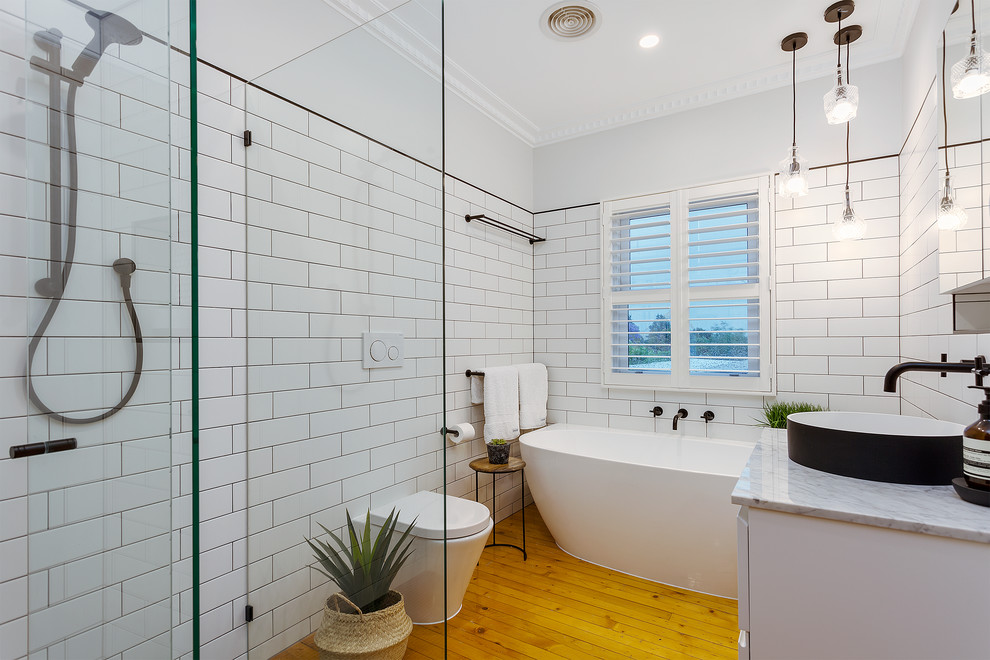 Modern inredning av ett vit vitt badrum, med släta luckor, vita skåp, ett fristående badkar, en öppen dusch, en vägghängd toalettstol, vit kakel, vita väggar, ljust trägolv, ett fristående handfat, brunt golv och med dusch som är öppen