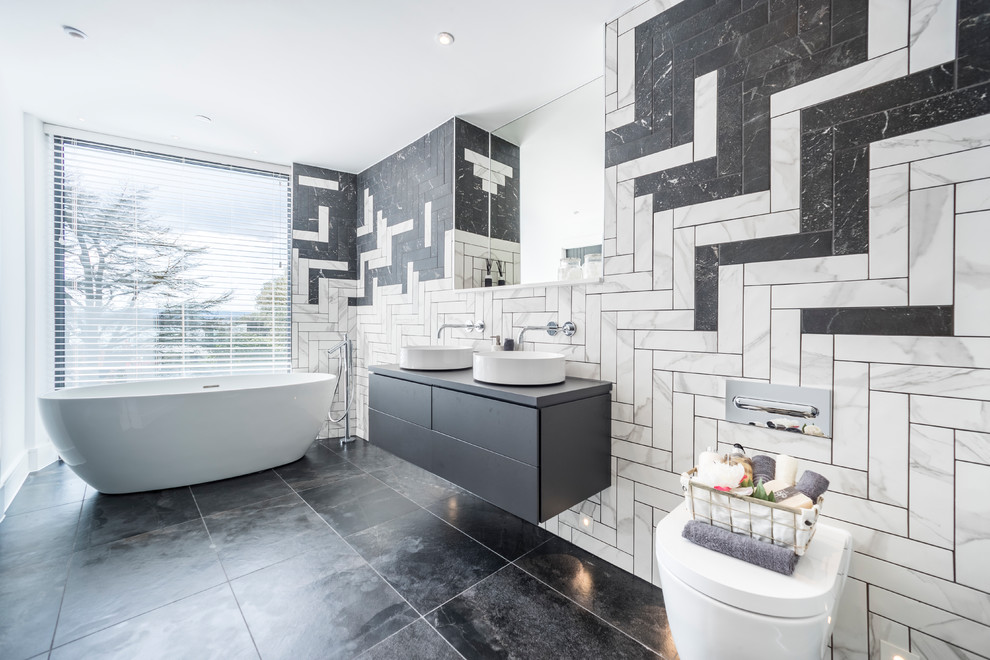 На фото: большая ванная комната в современном стиле с плоскими фасадами, серыми фасадами, отдельно стоящей ванной, инсталляцией, черно-белой плиткой, серой столешницей, белыми стенами, настольной раковиной и черным полом