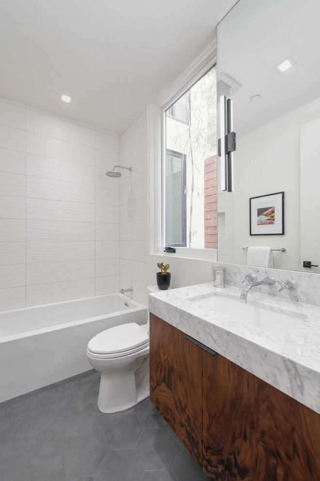 Bild på ett mellanstort funkis grå grått badrum, med släta luckor, skåp i mellenmörkt trä, ett badkar i en alkov, en dusch/badkar-kombination, en toalettstol med separat cisternkåpa, grå kakel, porslinskakel, grå väggar, ett undermonterad handfat, marmorbänkskiva och dusch med duschdraperi