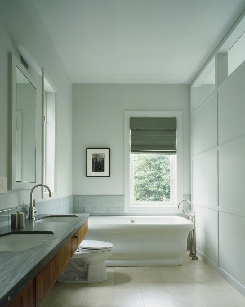 Inredning av ett klassiskt en-suite badrum, med ett fristående badkar, ett undermonterad handfat, släta luckor, skåp i mellenmörkt trä, en toalettstol med separat cisternkåpa, grå kakel, tunnelbanekakel, grå väggar, klinkergolv i porslin och grått golv