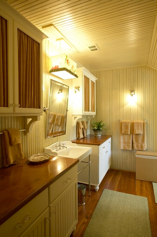 Modelo de cuarto de baño de estilo de casa de campo con lavabo con pedestal, armarios tipo mueble, puertas de armario blancas, paredes blancas y suelo de madera en tonos medios