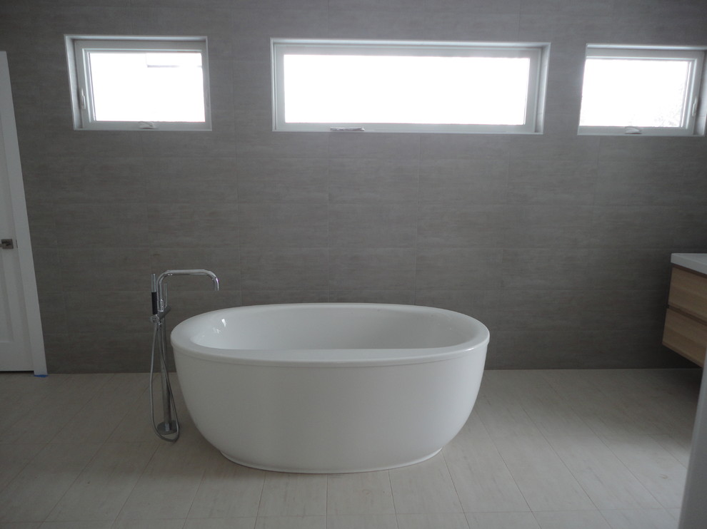 Cette photo montre une grande salle de bain principale moderne en bois clair avec un placard à porte plane, une baignoire indépendante, un carrelage gris, un mur gris, un lavabo intégré, un plan de toilette en quartz modifié, un sol beige et un plan de toilette blanc.