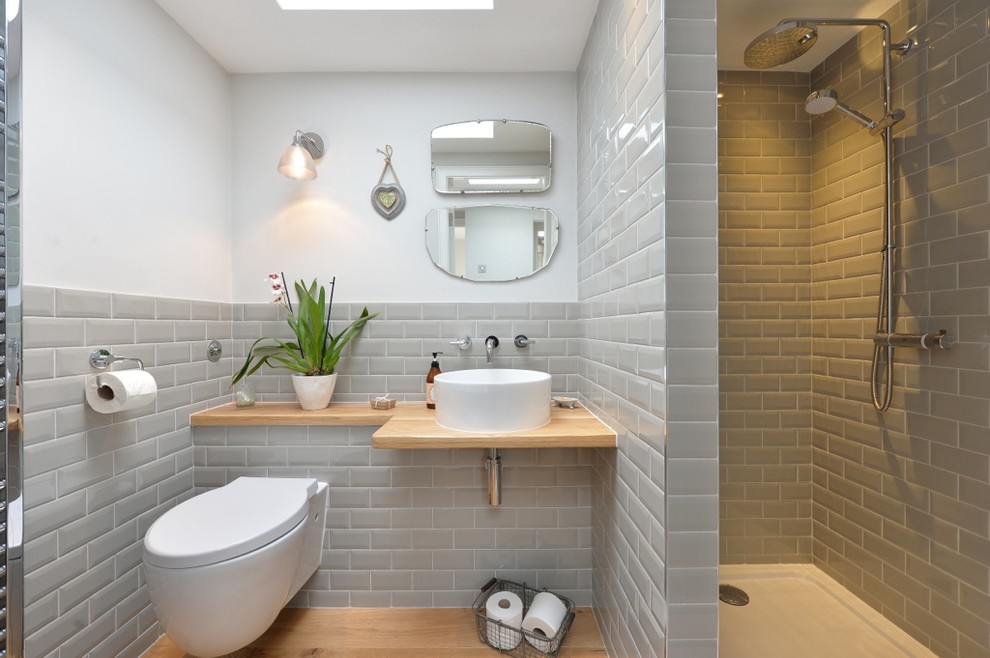 Foto på ett vintage beige badrum, med en öppen dusch, en vägghängd toalettstol, grå kakel, tunnelbanekakel, vita väggar, mellanmörkt trägolv, ett fristående handfat, träbänkskiva och med dusch som är öppen