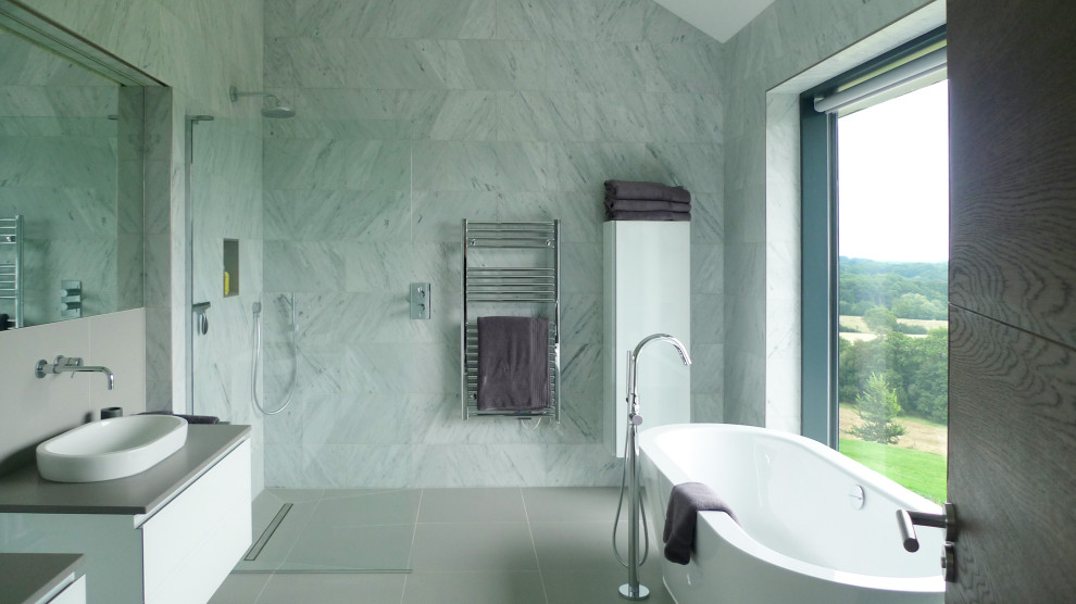 Modern inredning av ett grå grått badrum, med släta luckor, vita skåp, ett fristående badkar, grå kakel, ett fristående handfat och grått golv