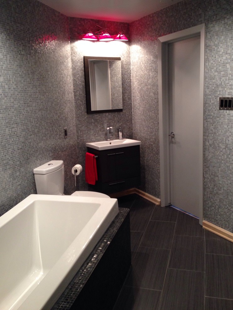 Modern inredning av ett en-suite badrum, med ett integrerad handfat, släta luckor, skåp i mörkt trä, ett platsbyggt badkar, en toalettstol med separat cisternkåpa, grå kakel, glaskakel och klinkergolv i keramik