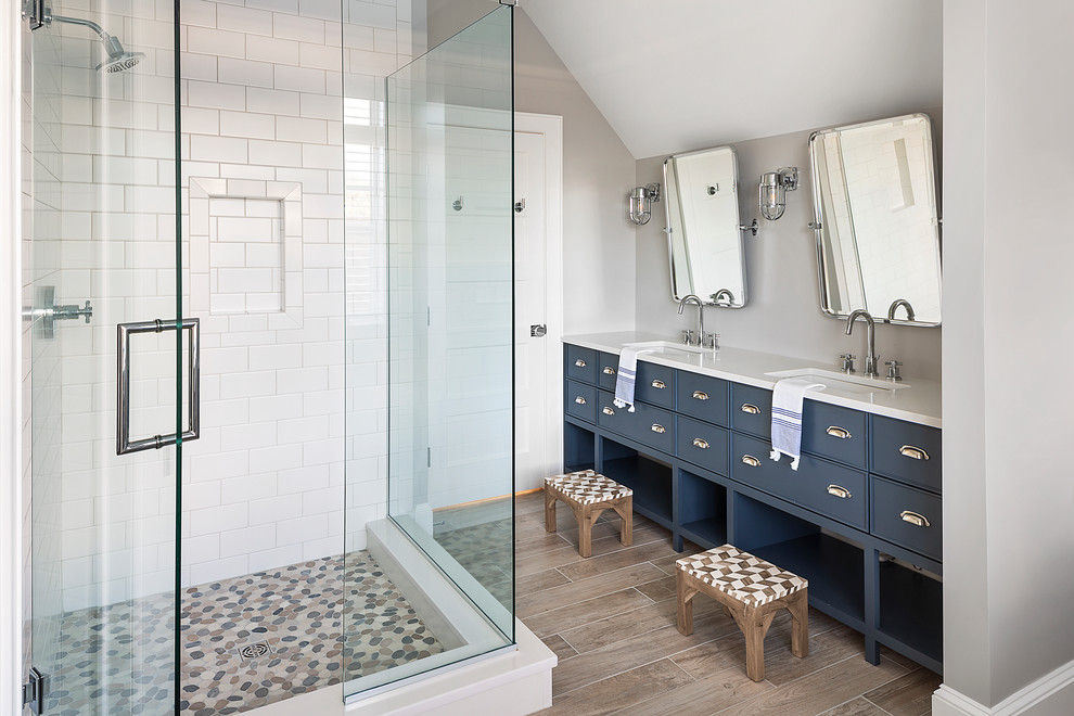 Cette image montre une salle de bain marine avec un placard à porte plane, des portes de placard bleues, une douche d'angle, un carrelage blanc, un mur gris, un lavabo encastré, un sol marron, une cabine de douche à porte battante et un plan de toilette blanc.