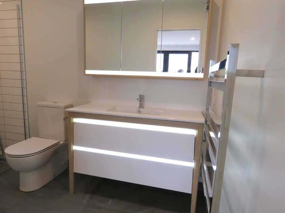 Inspiration for a medium sized modern ensuite bathroom in Dunedin with freestanding cabinets, white cabinets, a corner shower, a two-piece toilet, white tiles, white walls, a built-in sink, grey floors and white worktops.